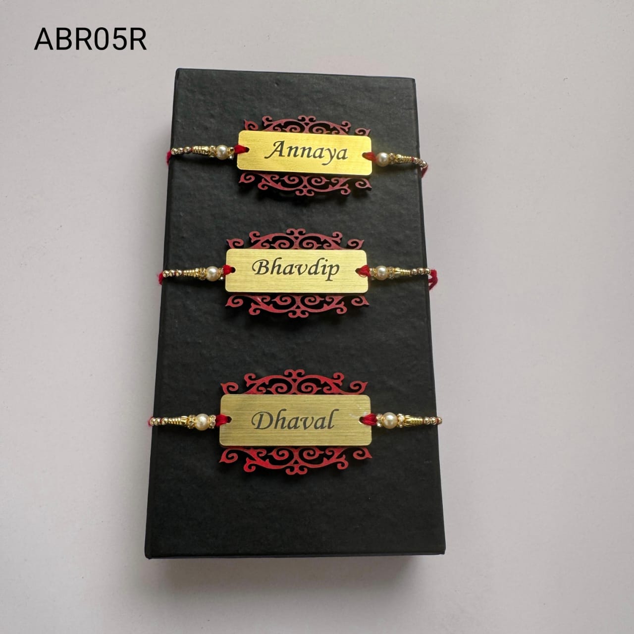 Personalized Gold Name Plate Designer Rakhi Personalized brushed gold name plate Rakhi with decorative designer base, pearl bead accents, and traditional red thread for Raksha Bandhan.